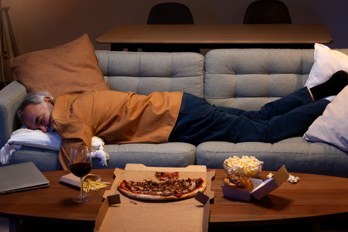 A person sleeping on a couch with unhealthy food on the table, symbolizing the effects of eating habits on sleep.