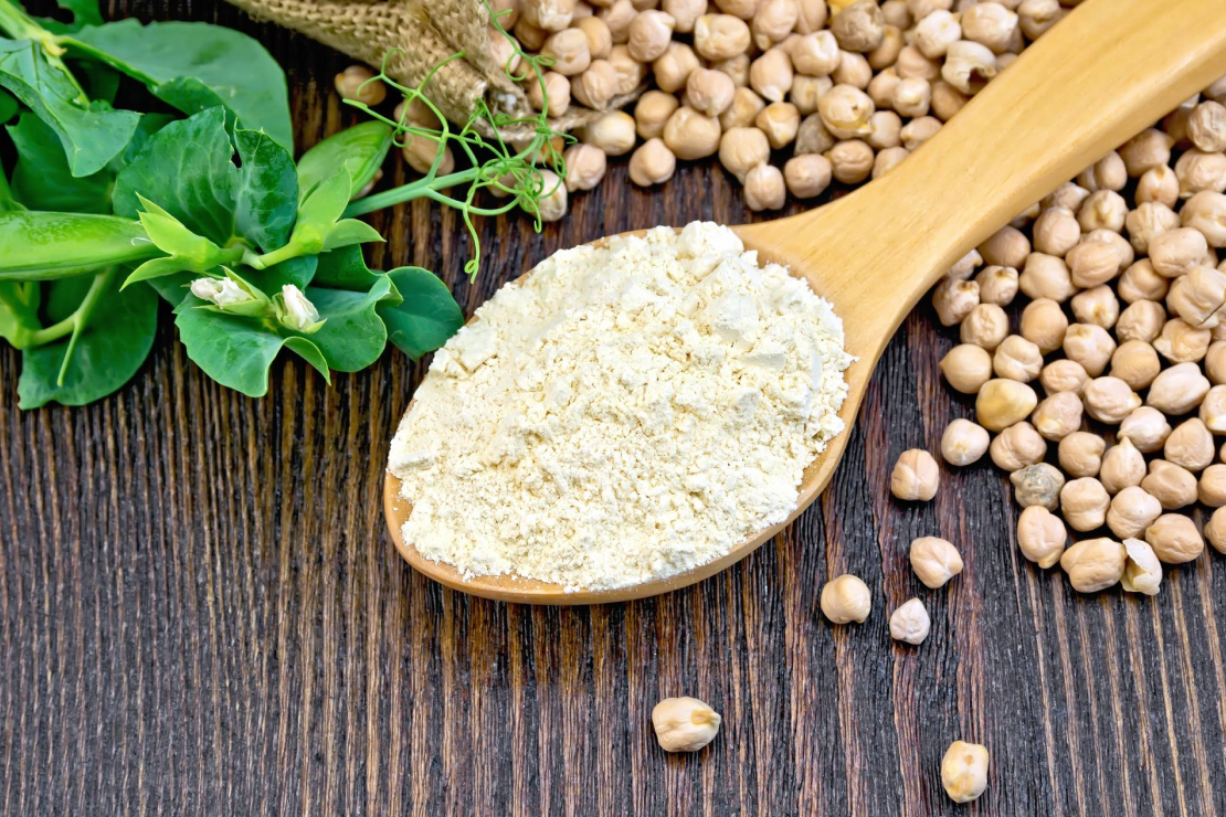 A wooden spoon with pea protein powder, surrounded by chickpeas and green pea pods, symbolizing plant-based protein.
