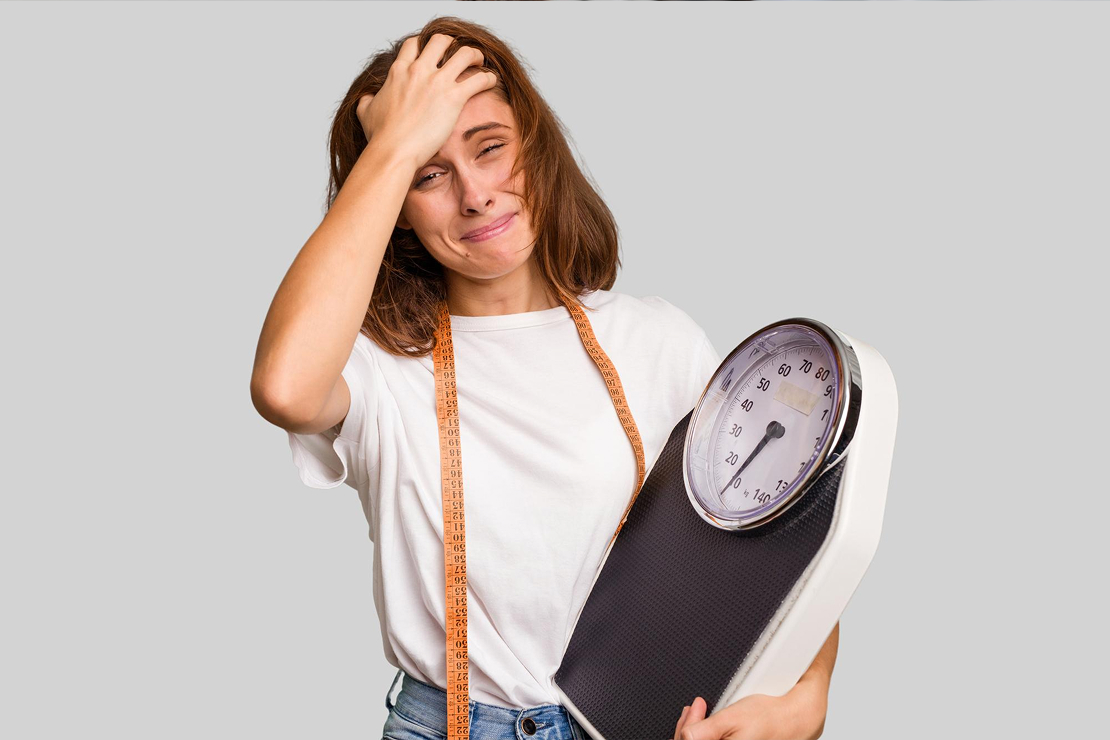 A woman looking distressed in front of a weight scale, symbolizing weight loss trauma.